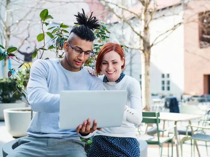 Two friends sit outdoors and look at a laptop together, exploring neighborhood businesses on a local directory